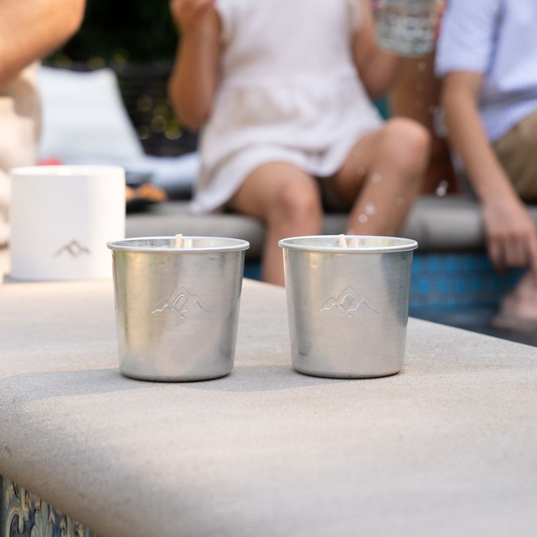 Two metal tin candle refills on a stone surface with blurred background depicting a gathering of people.