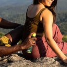 Two people sitting on the side of a mountain. The person behind  is spraying the other persons back of their arm