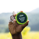 A hand holding a tin of mosquito repellent balm with a green lid, outdoors.