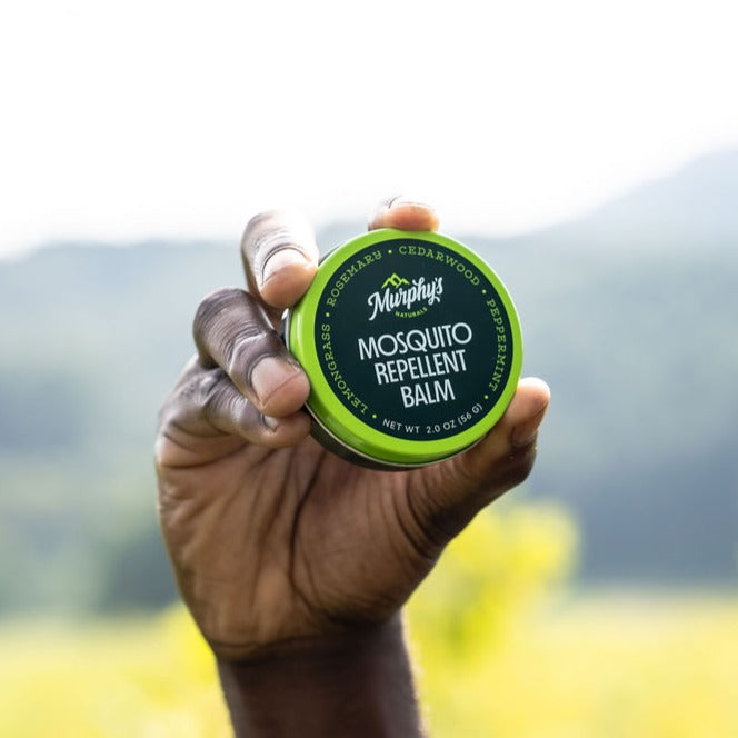 A hand holding a tin of mosquito repellent balm with a green lid, outdoors.
