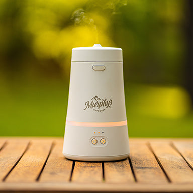 Murphy's Naturals Mosquito Dodger Zone Repellent device on a picnic table with a blurry green background. A fine mist is spraying from the top.