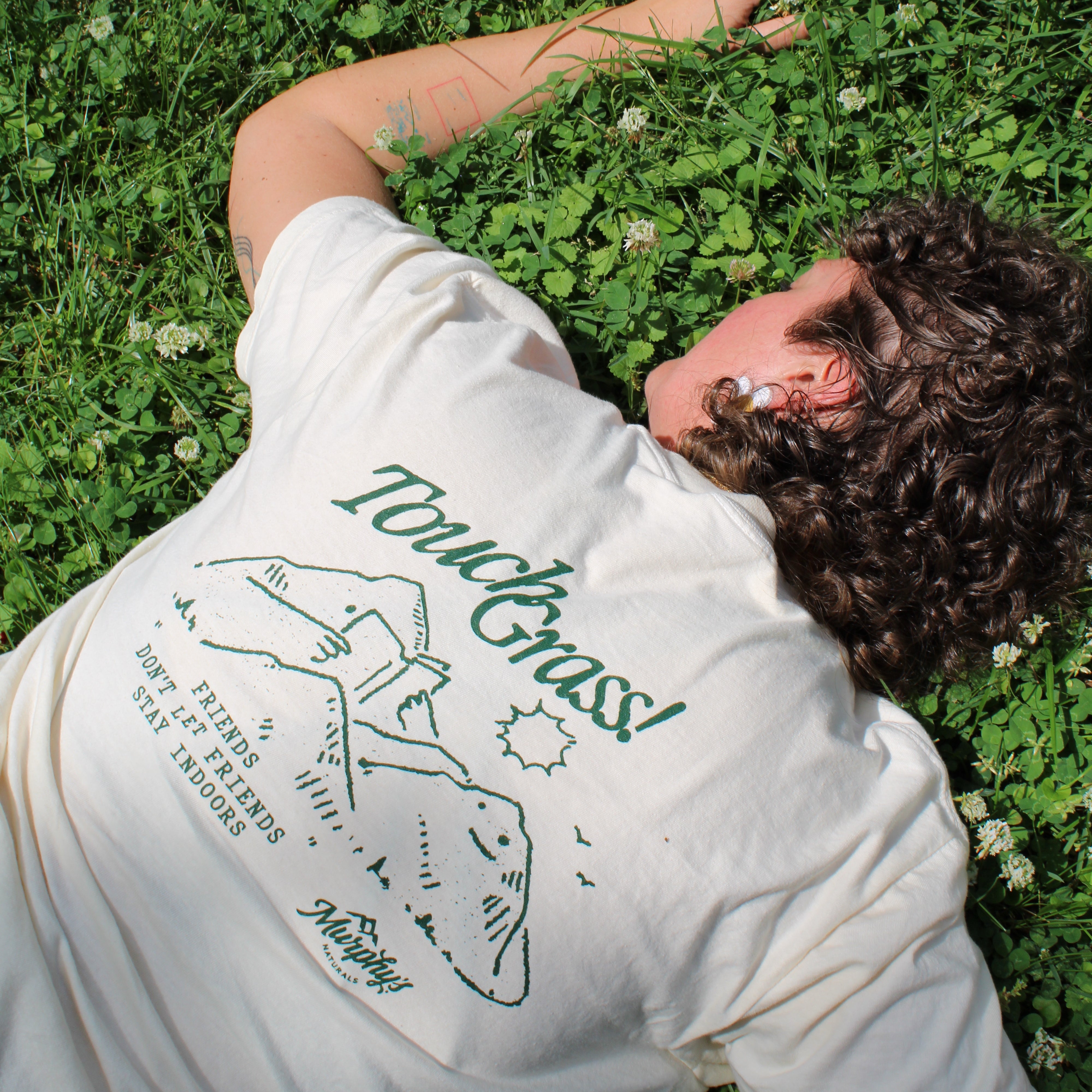 A person laying in the grass, the back of their shirt stating, "Touch grass" and depicting a mountain with a face reading a book.