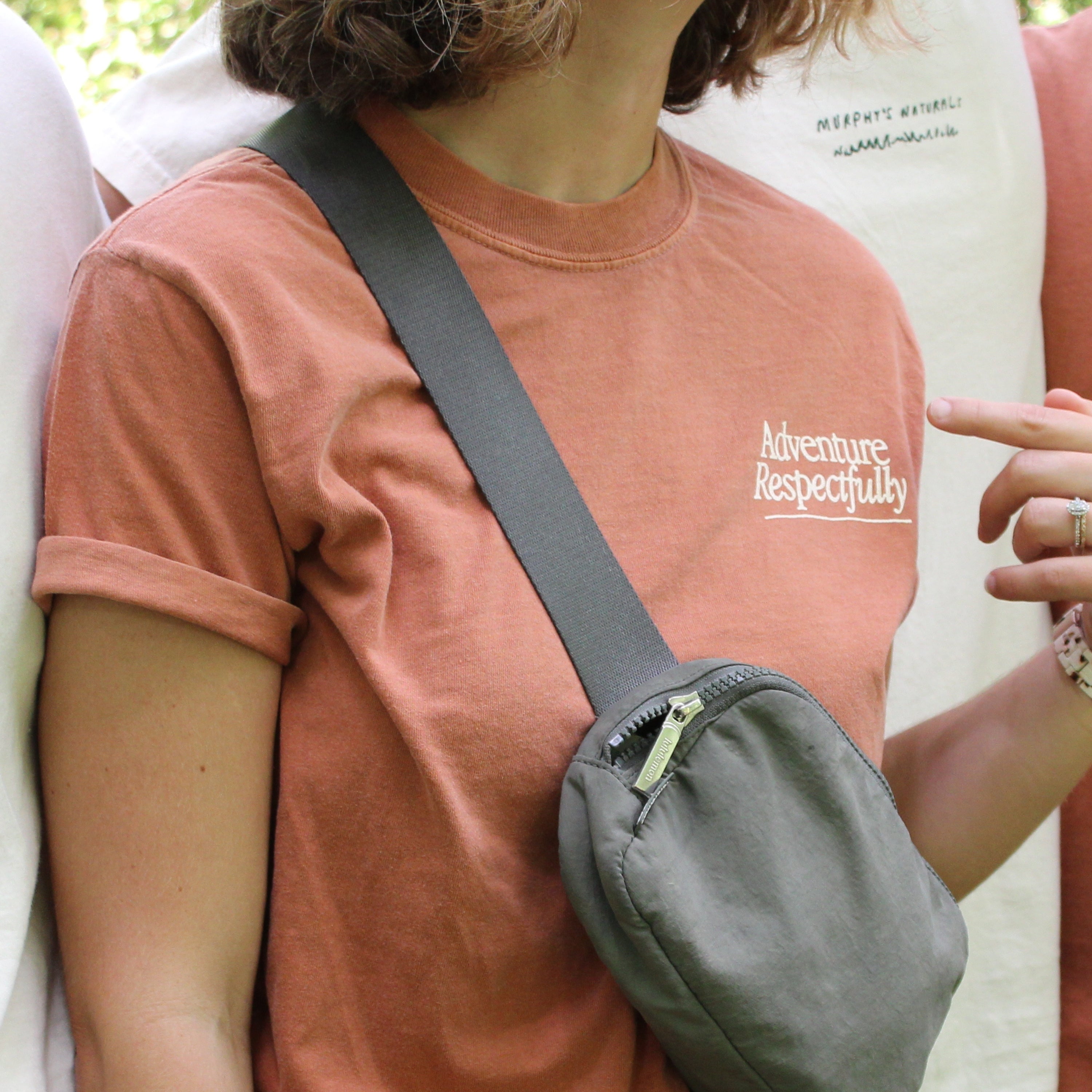 Adventure Respectfully on a t shirt. PErson wearing is pointing to it and wearing a bag slung over one shoulder.