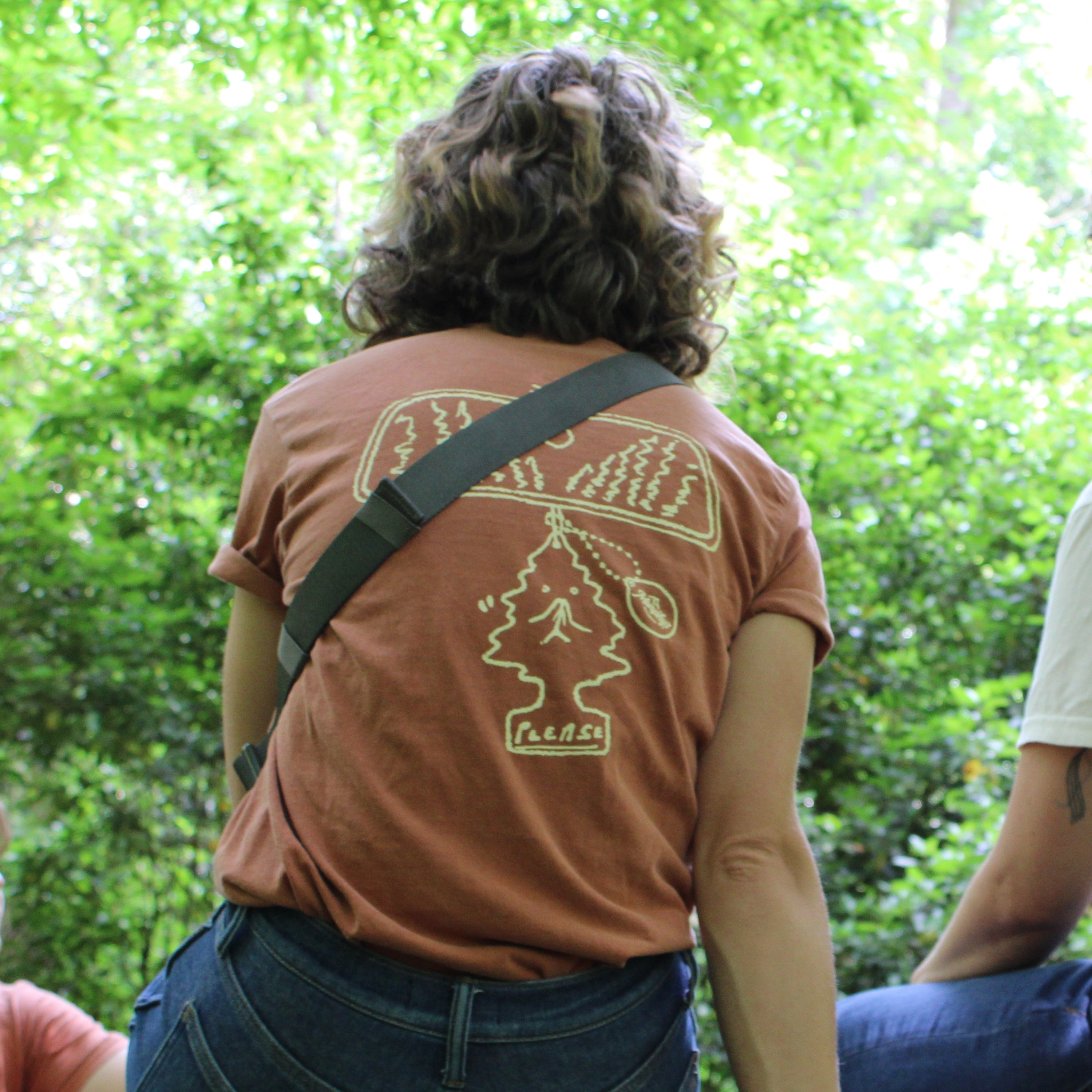 person sitting looking away wearing an orange shirt with a design epicting a rearview mirror with a scent tree hanging from it with "please" written at the bottom. Referencing the front of the shirt that says "adventure respectfully"