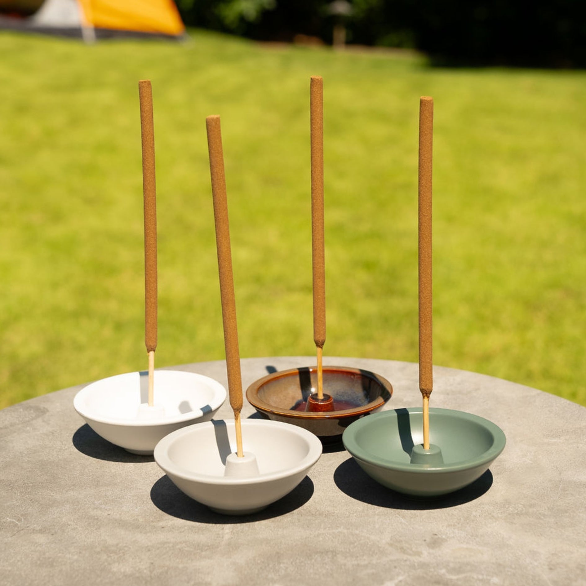 A collection of incense stick holders in white, brown, and green colors, each with a stick of incense placed in it, displayed outdoors on a flat surface.