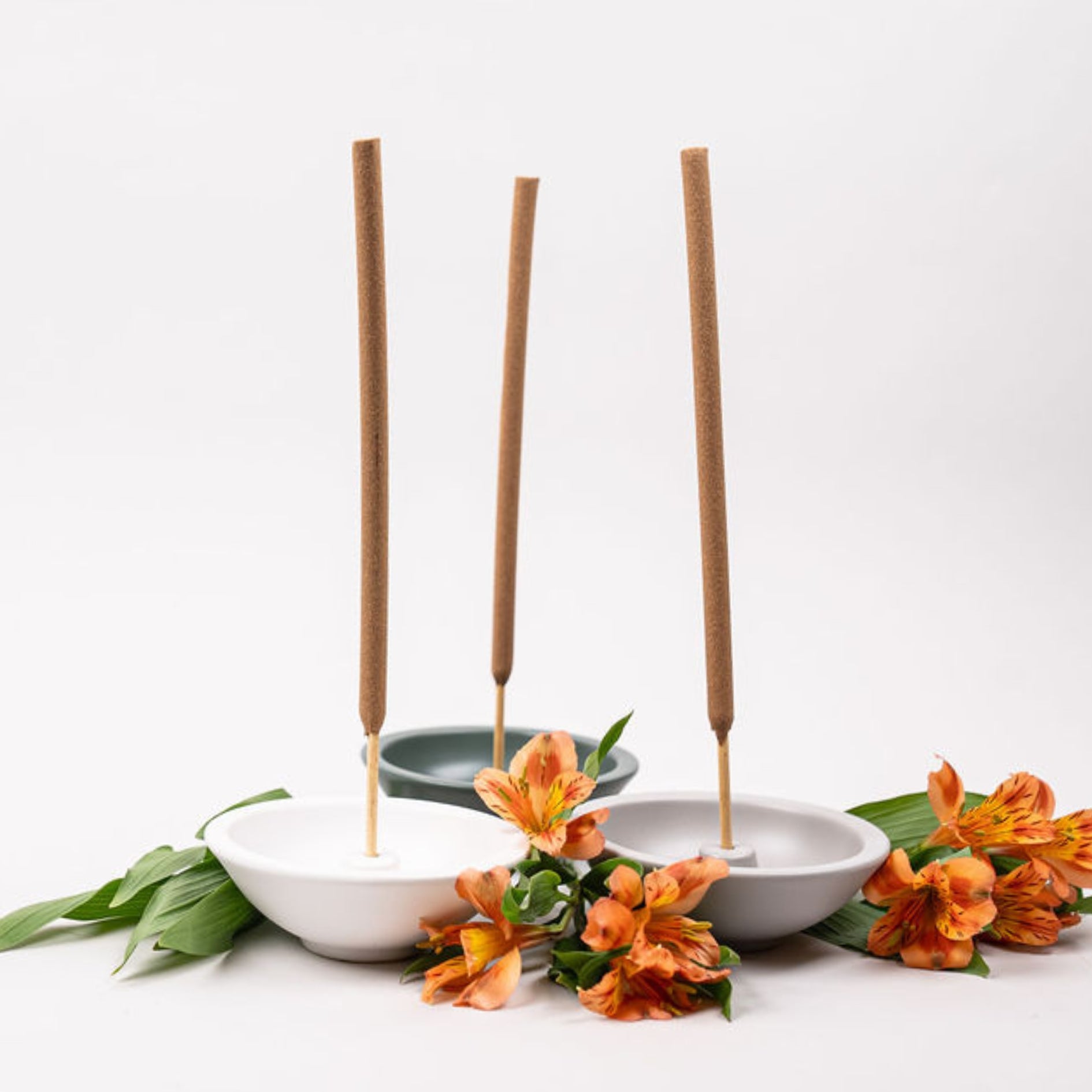 Incense stick holders in white, grey, and green with flowers around it on a white background