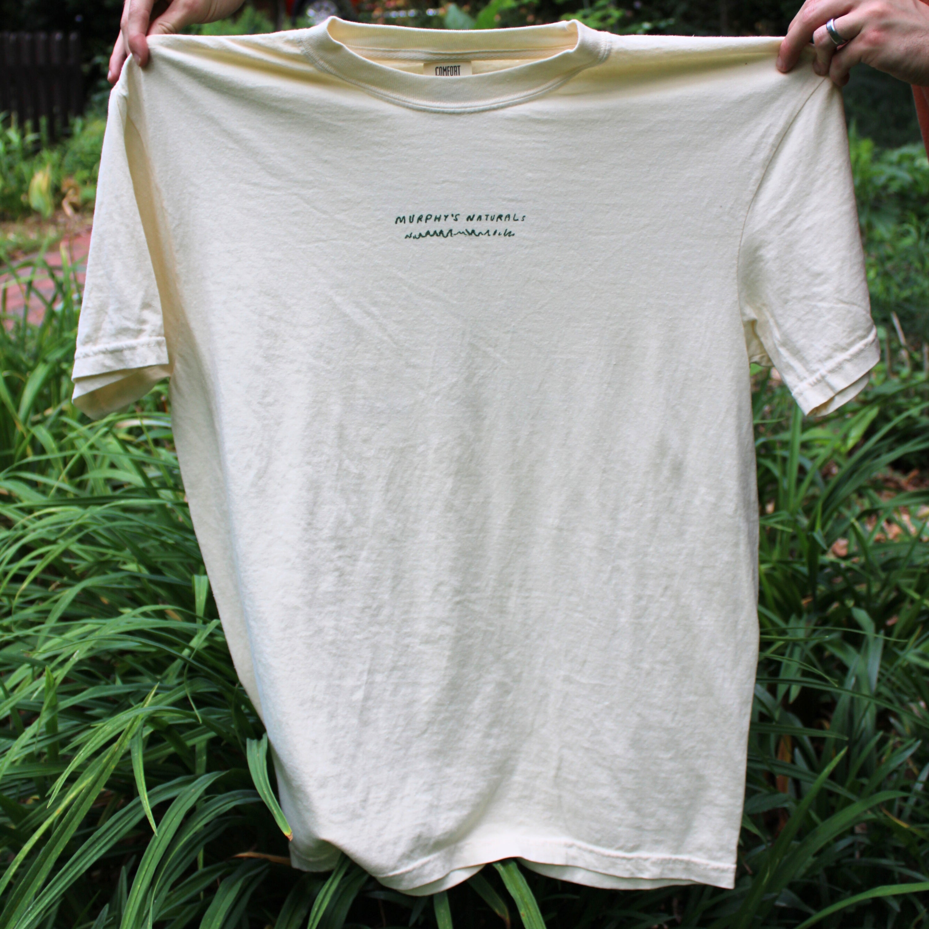 A cream t shirt with the text "Murphy's Naturals" held up in front of an outdoor scene.