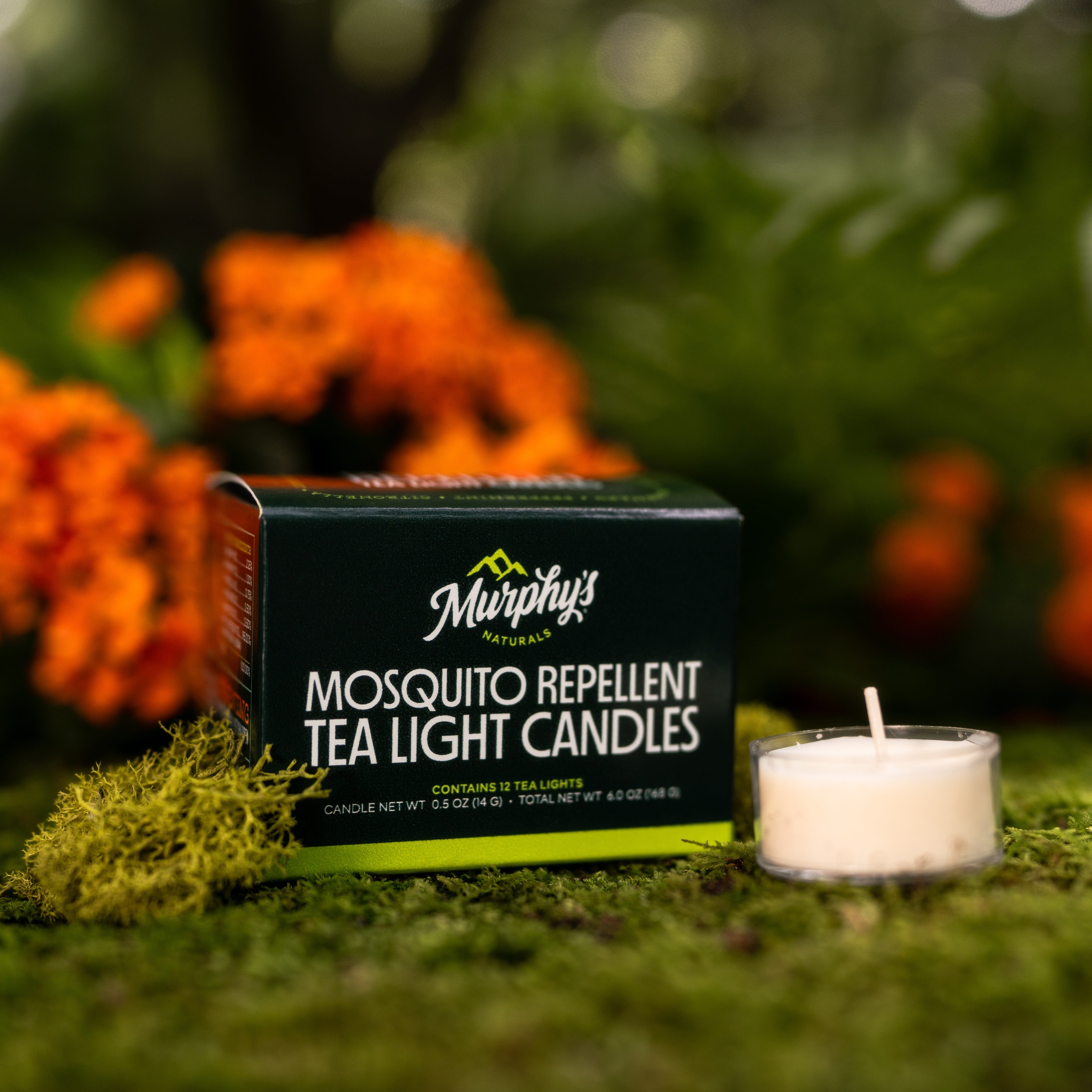 A mosquito repellent tea light candle from Murphy's Naturals is lit on a mossy surface with orange flowers in the background, indicating the product provides mosquito protection.