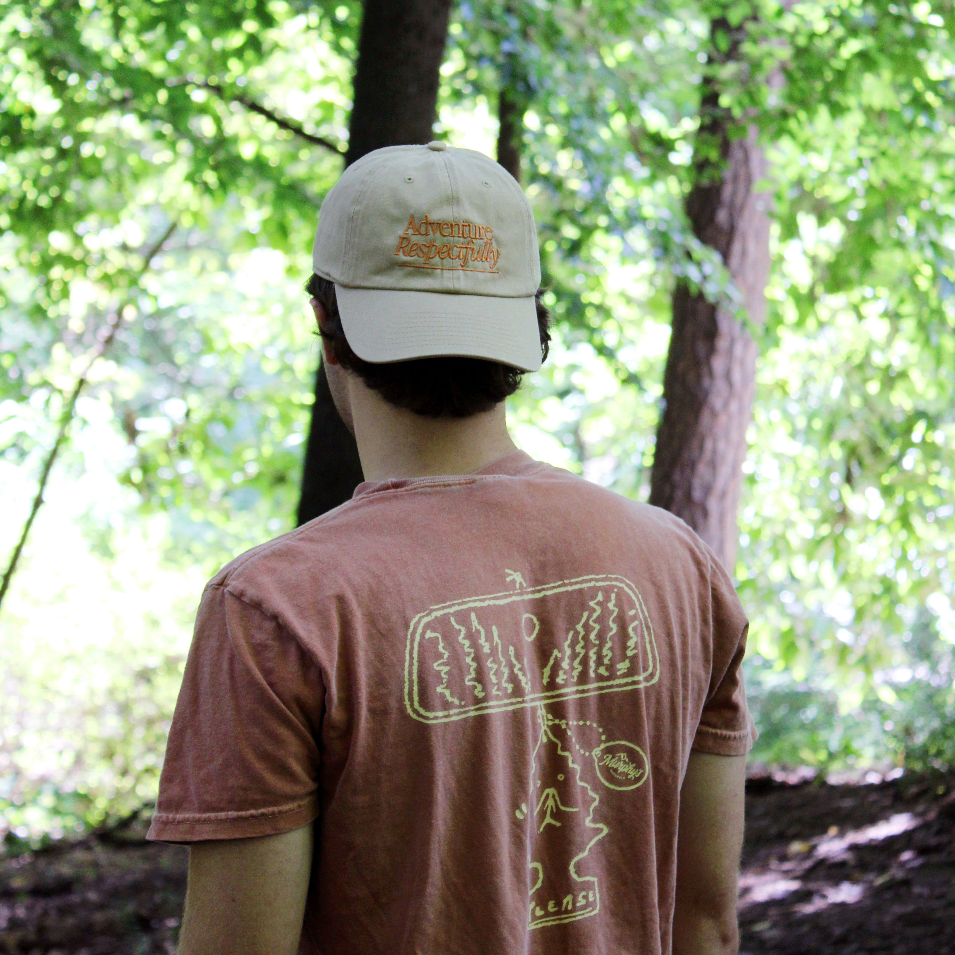 A person wearing a khaki-colored cap with 'ADVENTURE RESPECTFULLY.' embroidered on the front, standing in a wooded area.