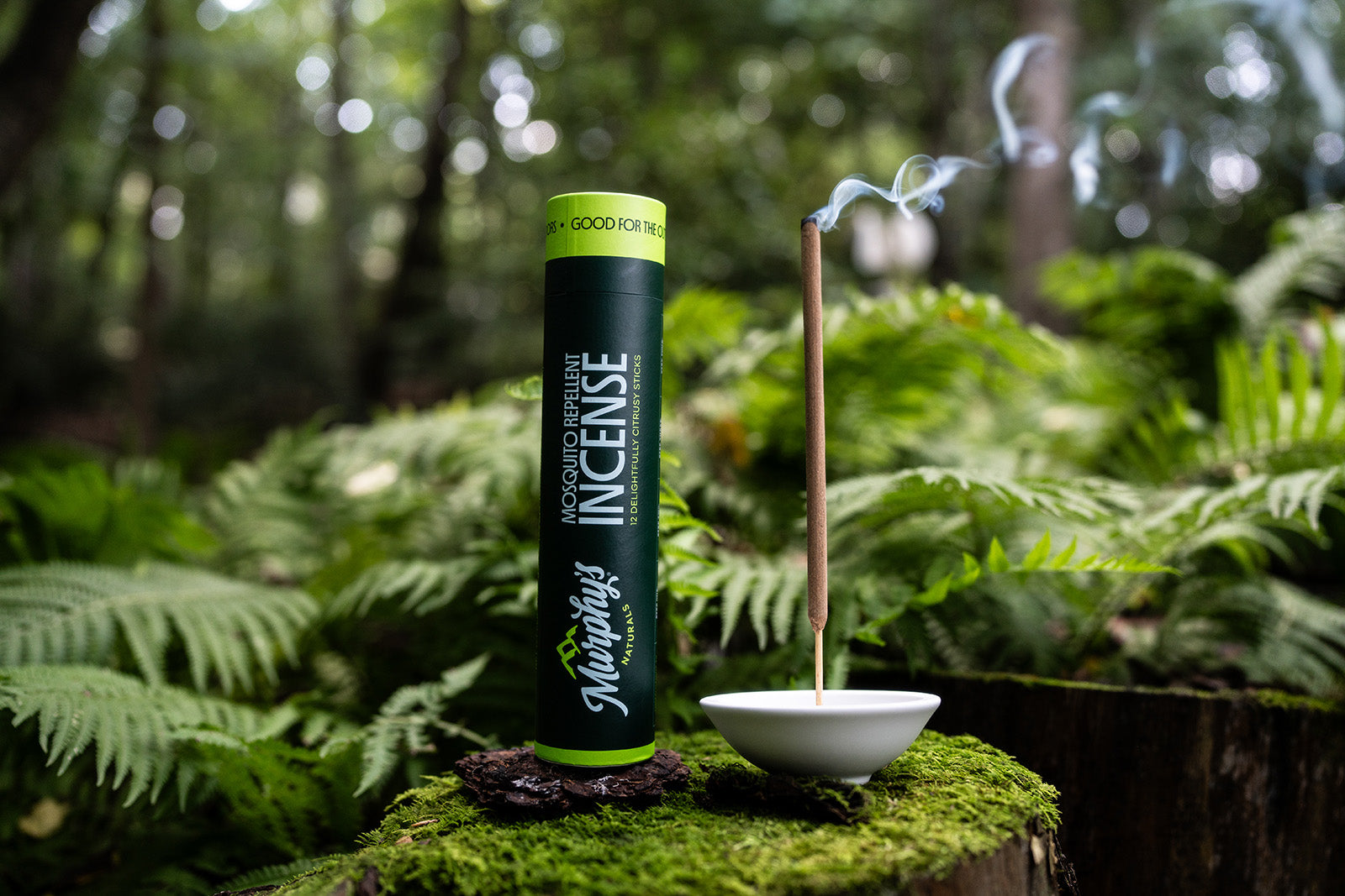 Murphy's Naturals Incense Stick with smoke coming from the burning stick. The Incense is sitting on a mossy stump and has ferns and greenery in the background.